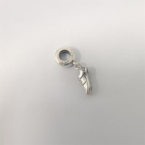 Könnte beinhalten: Ein silberner Charm mit einem kleinen Anhänger in Form eines Laufschuhs. Der Charm hat eine runde Schlaufe mit der Markierung "S 925" und einen kleinen Schuh mit detaillierten Merkmalen. Der Schuh ist als Laufschuh gestaltet.