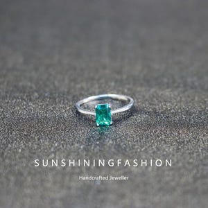 Peut inclure: Une bague en argent ornée d'une pierre précieuse rectangulaire vert émeraude. La bague est posée sur un fond sombre et texturé. Le texte "SUNSHININGFASHION" et "Handcrafted Jeweller" sont visibles.
