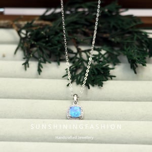 Puede incluir: Un collar de plata con un colgante cuadrado de ópalo azul claro. El colgante está rodeado de pequeñas piedras transparentes. El collar se muestra sobre una superficie gris claro con follaje verde en el fondo. El texto "SUNSHININGFASHION" y "Handcrafted Jewellery" son visibles.