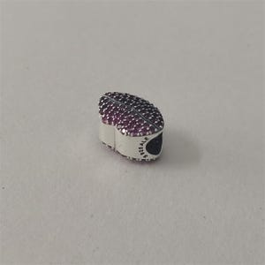 May include: A silver charm in the shape of a heart, adorned with numerous small, dark pink stones. The charm has a rounded top and a flat base. The side of the charm is marked with "S925 ALE".