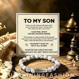 May include: A beaded bracelet with white and grey marbled stones and a rectangular silver-toned metal bar engraved with the name Thomas. Behind the bracelet is a square card with the printed text TO MY SON and a message about strength and support.