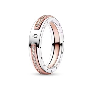 May include: A two-tone ring featuring a silver-colored band with the word PANDORA engraved along the side and a rose gold-colored inner band. The silver band includes a small crown logo and two rectangular sections inlaid with clear stones.