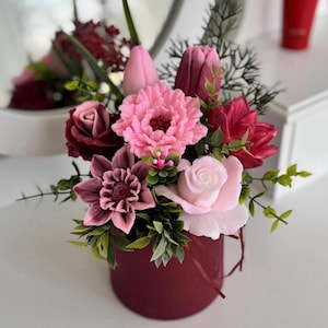 Peut inclure: Un arrangement floral dans une boîte ronde bordeaux. Il comprend des pivoines roses, des roses et des tulipes, avec des accents de feuillage vert. Les fleurs sont dans des tons de rose et de bordeaux.