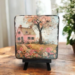 Peut inclure: Une peinture d'une maison rose avec un toit brun et un arbre avec des fleurs roses devant. La maison est entourée d'une clôture et de fleurs. La peinture est sur un petit chevalet.