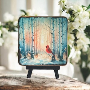 以下が含まれることがあります： 雪が降った白樺の森の中で、枝に止まっている赤いカージナルを描いた正方形の絵が載った小さな木製イーゼル。絵は青、白、ピンクの色合いで描かれている。