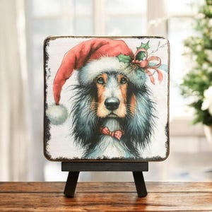 May include: A Christmas-themed wooden sign with a painting of a dog wearing a Santa hat and a red bow tie. The sign is on a small easel.