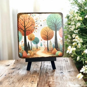Puede incluir: Un pequeño caballete de madera con una pintura cuadrada de una escena de bosque de fantasía. La pintura presenta árboles con hojas naranjas, amarillas y verdes, un camino de tierra y un pequeño pájaro volando en el cielo.
