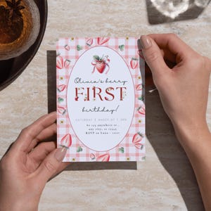 May include: A pink and white gingham patterned invitation with a red strawberry illustration. The text reads "Olivia's berry FIRST birthday!" with the date and time "SATURDAY | MARCH 27 | 2PM" and the location "At 123 anywhere st., any city, at 12345".