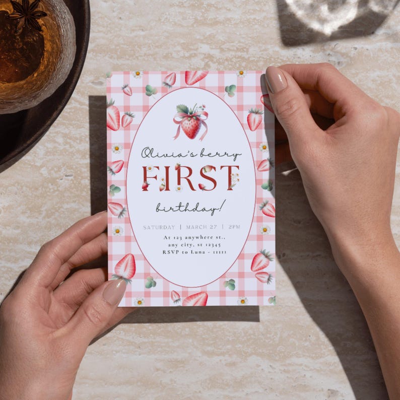 May include: A pink and white gingham patterned invitation with a red strawberry illustration. The text reads "Olivia's berry FIRST birthday!" with the date and time "SATURDAY | MARCH 27 | 2PM" and the location "At 123 anywhere st., any city, at 12345".
