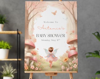 May include: A baby shower sign on a wooden easel. The sign features a whimsical illustration of a fairy in a forest setting with mushrooms and flowers. Text reads "Welcome to Artemis's Baby Shower Monday May 11th."