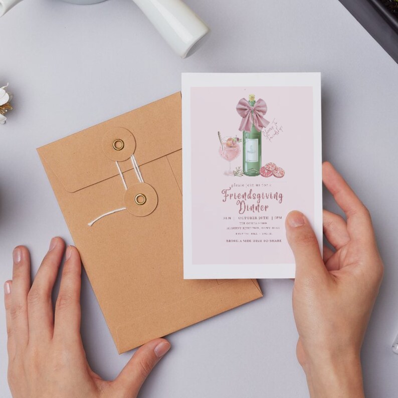 May include: A Friendsgiving Dinner invitation card held by a person, featuring a watercolor illustration of a bottle with a bow, a cocktail, and fruit. The invitation is on a pink background with the event details.