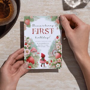 May include: A printable invitation for a first birthday party with a strawberry theme. The invitation features a girl in a red hooded dress walking through a strawberry patch with a mushroom and a bicycle in the background. The text on the invitation reads "Olivia's berry FIRST birthday!" and includes the date, time, and location of the party.
