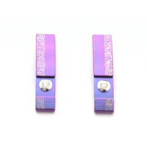 Könnte beinhalten: Ein Paar violette und blaue Ohrringe mit einem kleinen Diamanten in der Mitte jedes Ohrrings. Die Ohrringe haben ein dekoratives Muster an den Seiten.