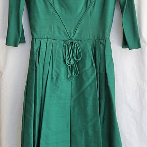 May include: Emerald green vintage dress with a square neckline, three-quarter sleeves, and a cinched waist. The dress features a V-shaped design on the bodice and a tie detail at the waist. The dress is approximately knee-length.