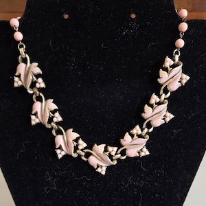 May include: A vintage necklace featuring pink leaf and berry designs. The leaves are a soft pink with gold-tone metal accents. Small white berry clusters are interspersed throughout the design. The necklace is displayed on a black surface.