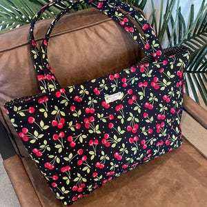 May include: A black tote bag with a cherry pattern. The bag features red cherries, green leaves, and white polka dots on a black background. The handles and interior lining are also black with a cherry pattern. The bag is sitting on a brown leather chair.