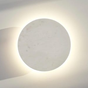 Puede incluir: Aplique de pared redondo y blanco con una luz suave y brillante. El aplique está montado en una pared blanca, proyectando una sombra a la izquierda. La luz ofrece un ambiente cálido y acogedor.