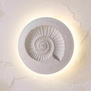 Può includere: Applique da parete rotonda bianca con un design a spirale fossile dettagliato. La luce emette un bagliore caldo, creando un effetto decorativo e ambientale. L'applique è montata su una parete bianca strutturata. Diametro circa 30 cm.