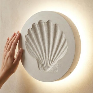 Puede incluir: Aplique de pared redondo blanco roto con un diseño detallado de concha. La concha está en relieve y texturizada, sobre un fondo circular. El aplique emite un brillo cálido, creando un ambiente costero o playero.