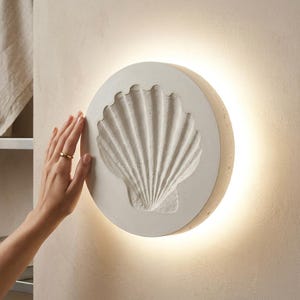 Op de afbeelding: Een ronde wandlamp met een getextureerd gebroken wit betonnen oppervlak en een verdiept reliëf van een sint-jakobsschelp in het midden. Het armatuur verspreidt een zachte, warme gloed vanaf de randen, waardoor de muur wordt verlicht.