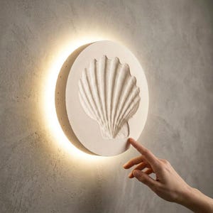 Puede incluir: Aplique de pared redondo, color crema, con diseño de concha marina en relieve. La luz emitida desde atrás crea un brillo cálido. La concha tiene una superficie texturizada y elevada. El aplique está montado en una pared gris.