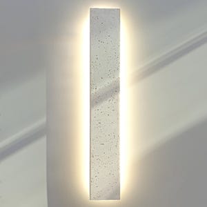 Puede incluir: Aplique de pared rectangular gris claro texturizado con una luz cálida y brillante que emana del centro. El aplique está montado en una pared blanca, creando una estética moderna y minimalista.