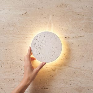 Puede incluir: Una lámpara de pared redonda, blanquecina, con una superficie texturizada y una luz cálida. La lámpara es sostenida por una mano contra una pared de piedra beige. Diseño moderno y minimalista para la decoración de interiores.