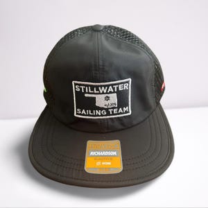 May include: Black baseball cap with a curved brim and mesh back. The cap features a white patch with the words "STILLWATER SAILING TEAM" and a logo. A yellow and orange tag is attached to the brim.