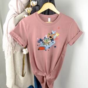 May include: A dusty rose t-shirt with a graphic print of Stitch, Baby Yoda, and Mickey Mouse riding in a car with balloons. The graphic is in a cartoon style.