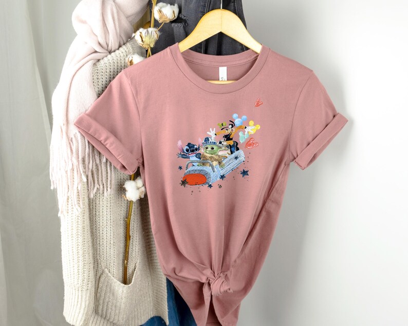 May include: A dusty rose t-shirt with a graphic print of Stitch, Baby Yoda, and Mickey Mouse riding in a car with balloons. The graphic is in a cartoon style.