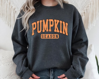 May include: Dark grey sweatshirt featuring the words "PUMPKIN SEASON" in orange block letters with a black outline. The crew neck sweatshirt is worn by a person. The text is in a collegiate style.