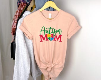 May include: A peach-colored t-shirt with rolled-up sleeves. The shirt features the words "Autism Mom" in green and red, with a colorful puzzle piece heart design. The shirt is knotted at the bottom.