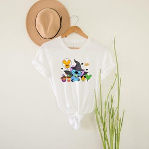 May include: White t-shirt featuring a blue Stitch character dressed as a witch with a black hat and holding a cauldron. The design also includes a yellow balloon, a purple ghost, and a green tree.