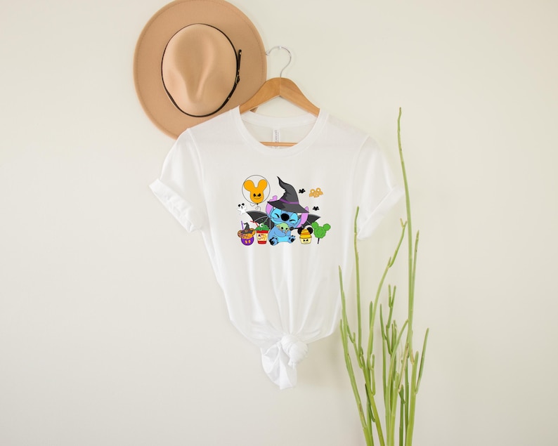 May include: White t-shirt featuring a blue Stitch character dressed as a witch with a black hat and holding a cauldron. The design also includes a yellow balloon, a purple ghost, and a green tree.