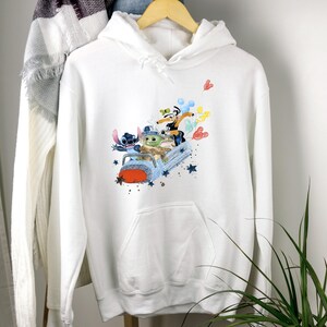 May include: White hooded sweatshirt with a colorful graphic print of Stitch, Baby Yoda, and Mickey Mouse riding in a spaceship. The graphic is surrounded by stars and hearts.