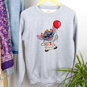 Pode incluir: Camisola de cor cinzenta com uma personagem Stitch azul vestida de palhaço a segurar um balão vermelho. A personagem tem um fato de palhaço branco e vermelho com um nariz vermelho e uma cara azul.