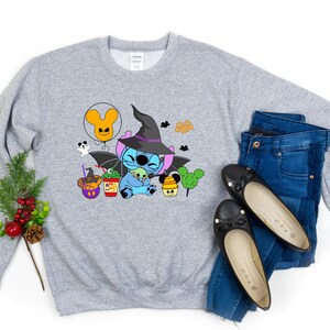 May include: A grey sweatshirt featuring a cartoon image of Stitch dressed as a witch, wearing a black hat and a purple dress. The design also includes a Mickey Mouse ear balloon, a cauldron, and a pumpkin. The sweatshirt is styled with blue jeans and black flats.