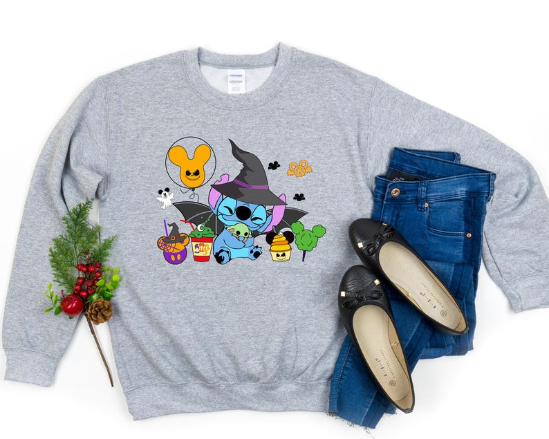 May include: A grey sweatshirt featuring a cartoon image of Stitch dressed as a witch, wearing a black hat and a purple dress. The design also includes a Mickey Mouse ear balloon, a cauldron, and a pumpkin. The sweatshirt is styled with blue jeans and black flats.