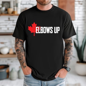 Puede incluir: Camiseta negra con una hoja de arce roja y el texto "Elbows Up" en blanco.