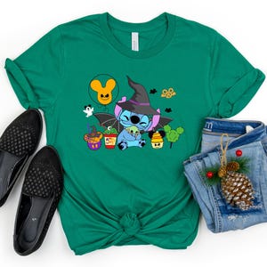 May include: Green t-shirt featuring a cartoon Stitch dressed as a witch, holding a smaller Stitch. The shirt has a Halloween theme with other cartoon characters and a Mickey Mouse balloon.