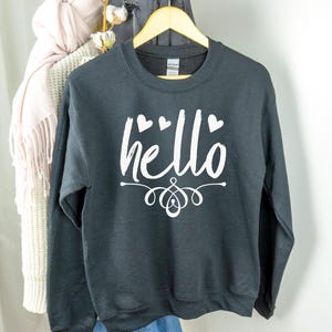 以下が含まれることがあります： 黒いスウェットシャツに、白い文字で「hello」と書いてあり、その周りにハートマークが描かれています。