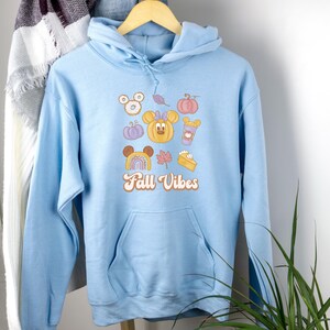 Pode incluir: Um moletom azul claro com um bolso frontal e capuz com cordão. A frente apresenta um gráfico colorido com imagens temáticas de outono, incluindo abóboras, folhas e o texto "Fall Vibes."
