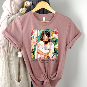 May include: A mauve t-shirt with a colorful illustration of Jesus holding a lamb. The text "I am the Resurrection & the Life John 11:25" is printed below the image.