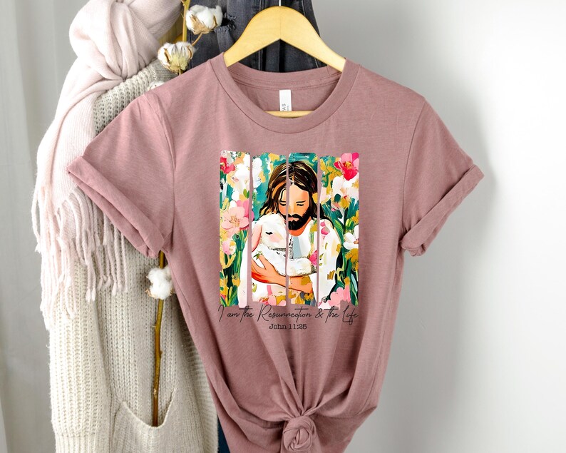 May include: A mauve t-shirt with a colorful illustration of Jesus holding a lamb. The text "I am the Resurrection & the Life John 11:25" is printed below the image.