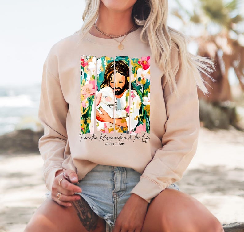 May include: A beige sweatshirt with a colorful illustration of Jesus holding a lamb. The illustration is surrounded by flowers and the text "I am the Resurrection & the Life John 11:25".