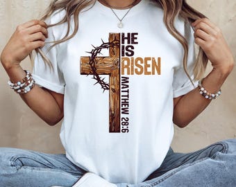May include: White t-shirt with a brown wooden cross and the text "He is Risen Matthew 28:6" printed on it.