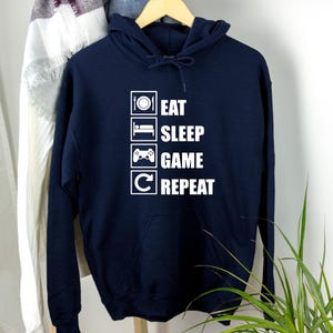 Könnte beinhalten: Ein marineblauer Hoodie mit einer weißen Grafik, auf der "Essen Schlafen Spielen Wiederholen" in einer Checklistenform steht.
