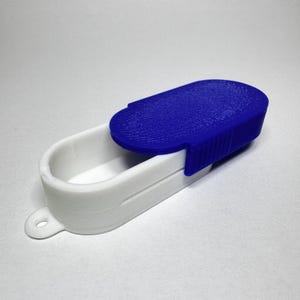 May include: A white and blue pill case with a textured blue lid. The case is oval-shaped with a small loop on one end. The lid is hinged and opens to reveal the interior of the case.