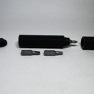 Op de afbeelding: Een zwarte metalen schroevendraaierhandgreep met een getextureerde ruitpatroon-grip, voorzien van een verwijderbare eindkap en een holle binnenkant. De set bevat twee verwisselbare stalen schroevendraaierbits, een platte en een kruiskop, en een dop.