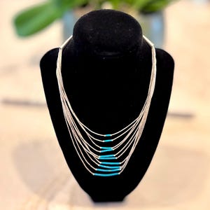 Puede incluir: Un collar de plata con múltiples cadenas y detalles turquesa. El collar se muestra en un maniquí de busto negro.
